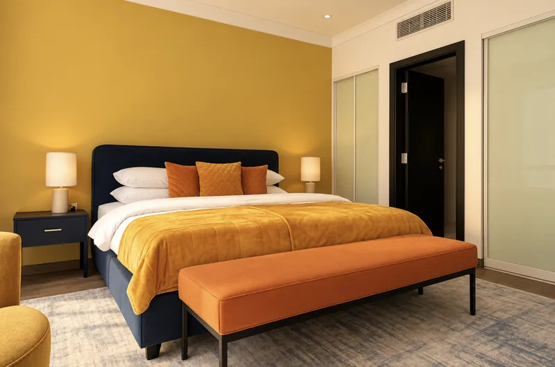 Dunia bedroom with mustard-yellow feature wall, navy bed, ochre throw, orange bench and built-in wardrobes