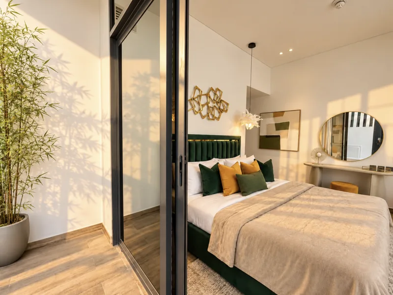 Pearl emerald-green bedroom seen through open terrace doors, with vanity, circular mirror and mustard ottoman
